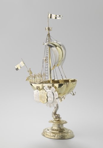 Rederijbeker van verguld zilver in de vorm van een schip gedragen door een dolfijn. Het schip is getuigd met mast, want en zeil en is bemand met 12 man.